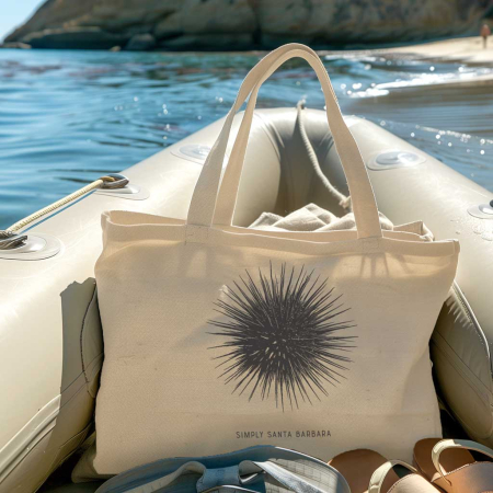 A natural fabric tote bag rests on an inflatable boat with the ocean in the background. The bag features a black sea urchin print with the 'Simply Santa Barbara' logo, capturing the essence of a day out at sea and the connection with marine life.