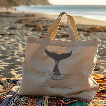 An eco-friendly tote bag resting on a colorful beach blanket on the sandy shore. The bag has a serene whale tail print emerging from the water with 'Simply Santa Barbara' written beneath. The late afternoon sun casts a gentle light, highlighting the peaceful beach setting.
