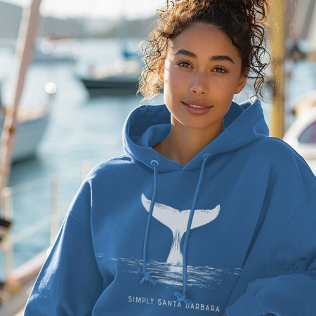 A young woman with a serene expression, wearing a indigo blue hoodie with a large off-white print of a whale's tail above the water, and the words 'Simply Santa Barbara' below it. The hoodie reflects the oceanic theme, aligning with the brand's celebration of marine life and the coastal vibe of Santa Barbara. The marina in the background adds to the nautical atmosphere of the image.
