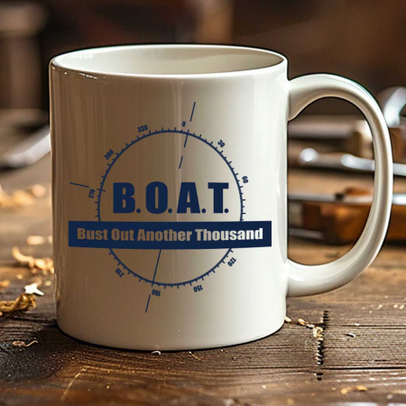 A white coffee mug with the humorous nautical acronym 'B.O.A.T.', standing for 'Bust Out Another Thousand', printed in blue letters along with a compass design, rests on a well-used wooden workbench scattered with wood shavings and tools. This image suggests the mug is a favored item among shipwrights or boat enthusiasts, capturing a moment of relaxation in a busy workshop, embodying the blend of craftsmanship and the boating lifestyle promoted by Simply Santa Barbara.