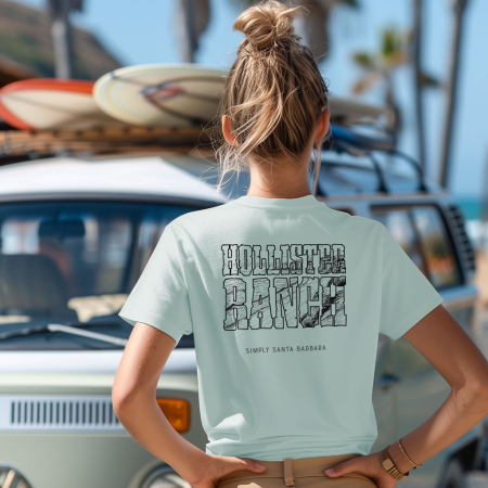 A woman stands with her back to the camera, hands on her hips, wearing a light blue green short-sleeve t-shirt with 'HOLLISTER RANCH' in a stylized black font on the back. Below the text is 'Simply Santa Barbara', indicating the shirt's local inspiration. In the background, there’s a vintage VW Bus with surfboards on top, parked by a palm-lined beach, suggesting a surfing or beach adventure.