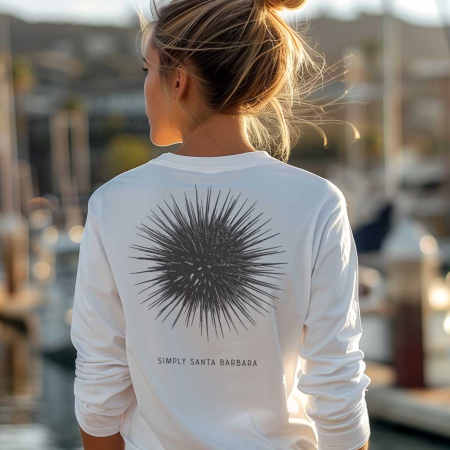 Viewed from behind, a woman with her hair styled in a high bun is wearing a white long-sleeve fitted crew shirt that features a large black sea urchin print on the back. Below the print, 'Simply Santa Barbara' is neatly scripted. She's standing at a marina with sunlit boats and calm waters in the background, evoking an elegant nautical vibe.