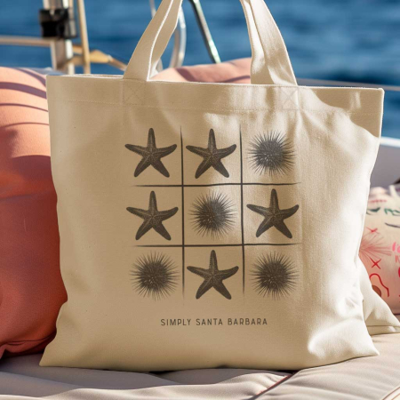 A natural beige organic tote bag with a black tic-tac-toe print featuring starfish and sea urchins as the game pieces. Below the game grid, the text 'Simply Santa Barbara' is displayed. The tote bag is propped against a cushion on a boat with the blue ocean in the background, suggesting a perfect accessory for a day of sailing or beach exploration.