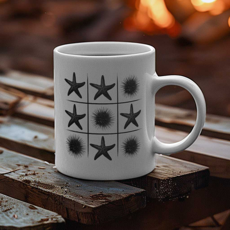 A white ceramic mug sits on a camp table, with an illustration of multiple black starfish and a sea urchin printed in a tic-tac-toe pattern on the mug. The mug's curved handle extends to the right side of the image, and the background shows a blurred, campfire on a beach.