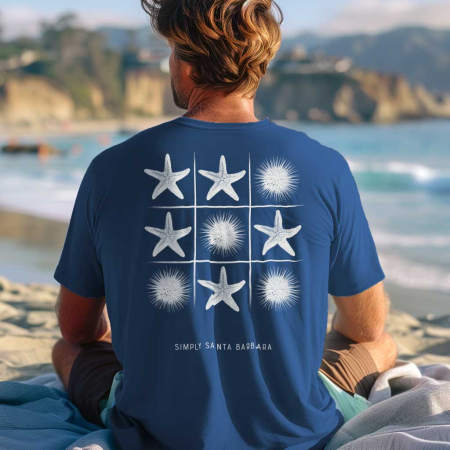 A man is seen from behind, sitting on a beach and looking out to the sea. He’s wearing a royal blue short-sleeve t-shirt with a tic-tac-toe grid on the back featuring white starfish and sea urchin designs as the game pieces. 'Simply Santa Barbara' is printed beneath the game. The tranquil beach and soft evening light create a peaceful, coastal setting