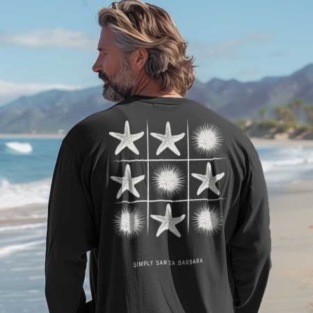 A man seen from behind with medium-length hair gazes towards the sea. He is wearing a heavy metal gray long-sleeve fitted crew shirt with a tic-tac-toe grid on the back featuring white starfish and sea urchins as pieces. The text 'Simply Santa Barbara' is printed below the game. The backdrop is a scenic beach with waves and distant mountains, suggesting a tranquil seaside setting.