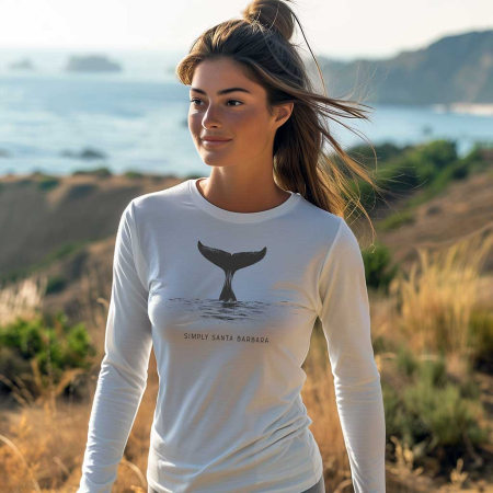 A woman with wind-tousled hair, facing sideways, wears a white long-sleeve fitted crew shirt featuring a centered black print of a whale's tail above the water. Below the print, 'Simply Santa Barbara' is printed. The setting is outdoors with coastal cliffs and a calm sea in the background, suggesting a connection with nature and marine life.