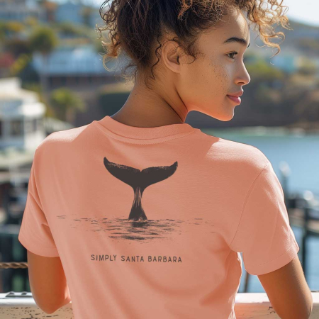 A woman with curly hair is viewed from the back, wearing a desert pink colored short-sleeve t-shirt that features a large black print of a whale tail diving into water, symbolizing connection to the ocean. The text 'Simply Santa Barbara' is printed beneath the image. She overlooks a harbor with boats and clear blue skies.