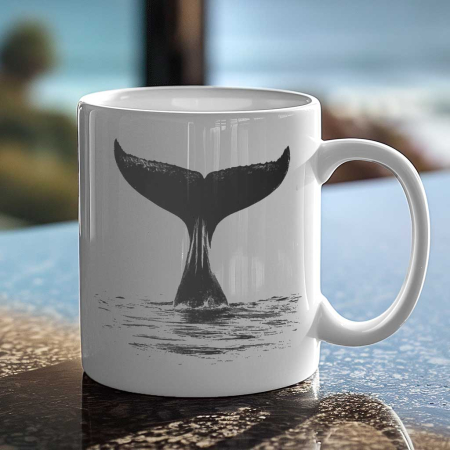 A glossy white mug with a graphic of a whale’s tail submerged in water, centered on the side facing the viewer. The mug rests on black granite countertop with a blurred coastal scene in the background, suggesting a peaceful morning coffee with an ocean view.