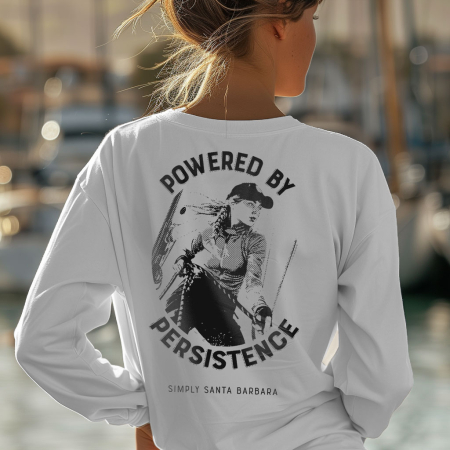 A woman from behind at the marina, hands on hips, wears a classic white long sleeve t-shirt with 'POWERED BY PERSISTENCE' printed in a bold font above a dynamic large dark gray graphic of a female sailor tailing sheets on a sailboat. The design signifies determination and the shirt boasts 'Simply Santa Barbara' underneath, indicating the brand. This high-quality shirt features a combed ringspun cotton build, a fitted design with a baby rib collar, suggesting comfort and style for a relaxed coastal lifestyle.