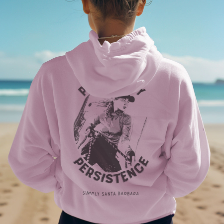 A woman on the beach is viewed from behind, wearing a soft lavender hoodie with the bold statement 'POWERED BY PERSISTENCE' and an image of a female sailor trimming the sheets on a sailboat printed on the back. The relaxed fit and the serene beach setting encapsulate a laid-back, inspirational coastal vibe."