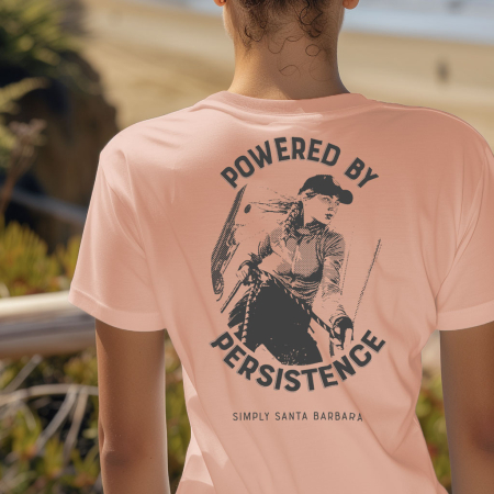 A woman is seen from behind, wearing a soft desert pink-colored short-sleeve t-shirt with a large dark gray graphic of a female sailor tailing sheets on a sailboat with the motivational phrase 'POWERED BY PERSISTENCE' arching over the image. Beneath the sailor, 'Simply Santa Barbara' is printed, linking the shirt to the coastal city's lifestyle. The setting is outdoors with sand dunes in the soft-focus background.